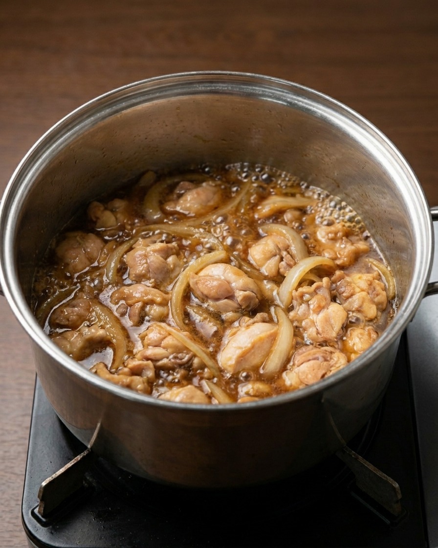 A pot on a stove with chicken pieces and onion slices simmering in a bubbling, dark brown savory liquid.