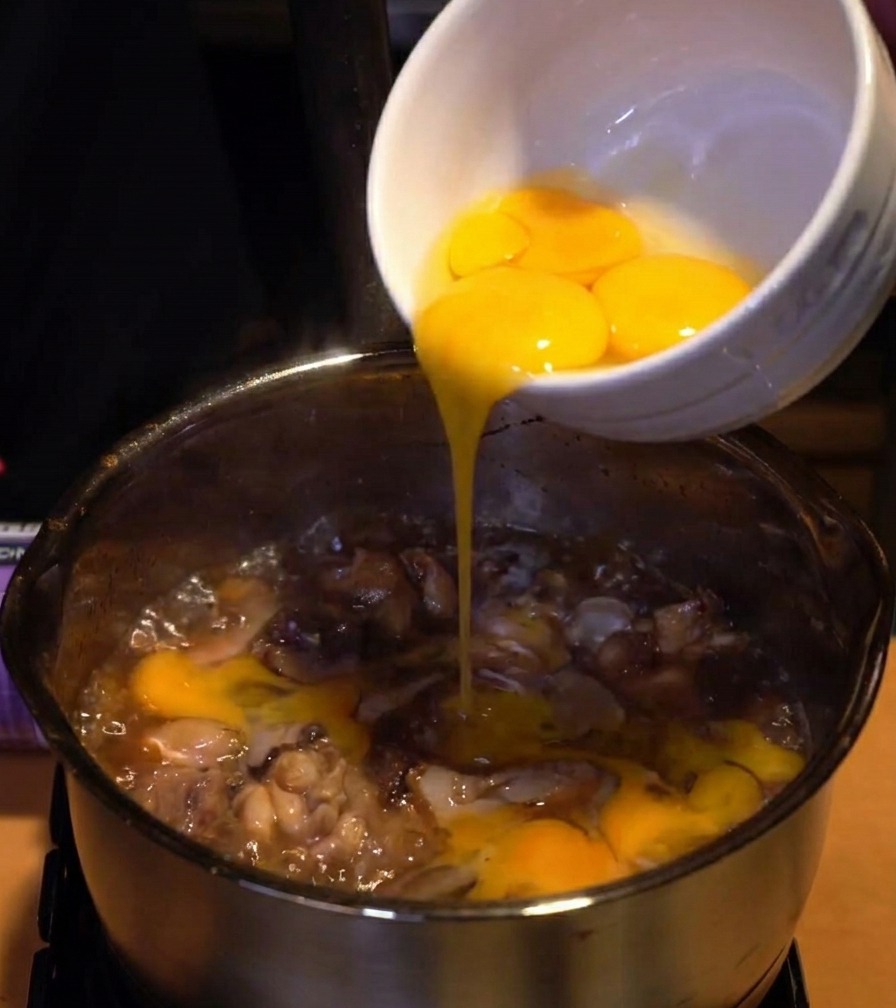 Egg whites being poured from a white ceramic bowl into a pot of simmering chicken and onions in a dark sauce.