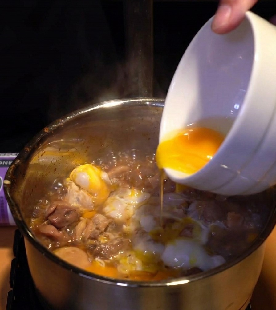 Vibrant yellow egg yolks being drizzled from a bowl over a layer of cooked white eggs and chicken in a simmering pot.