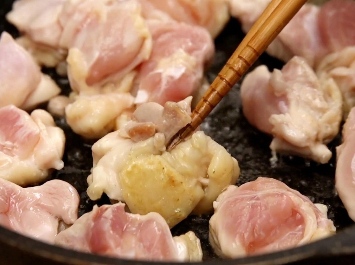Bite-sized chicken thigh pieces being sautéed in a dark frying pan with wooden chopsticks, showing some pieces beginning to brown.
