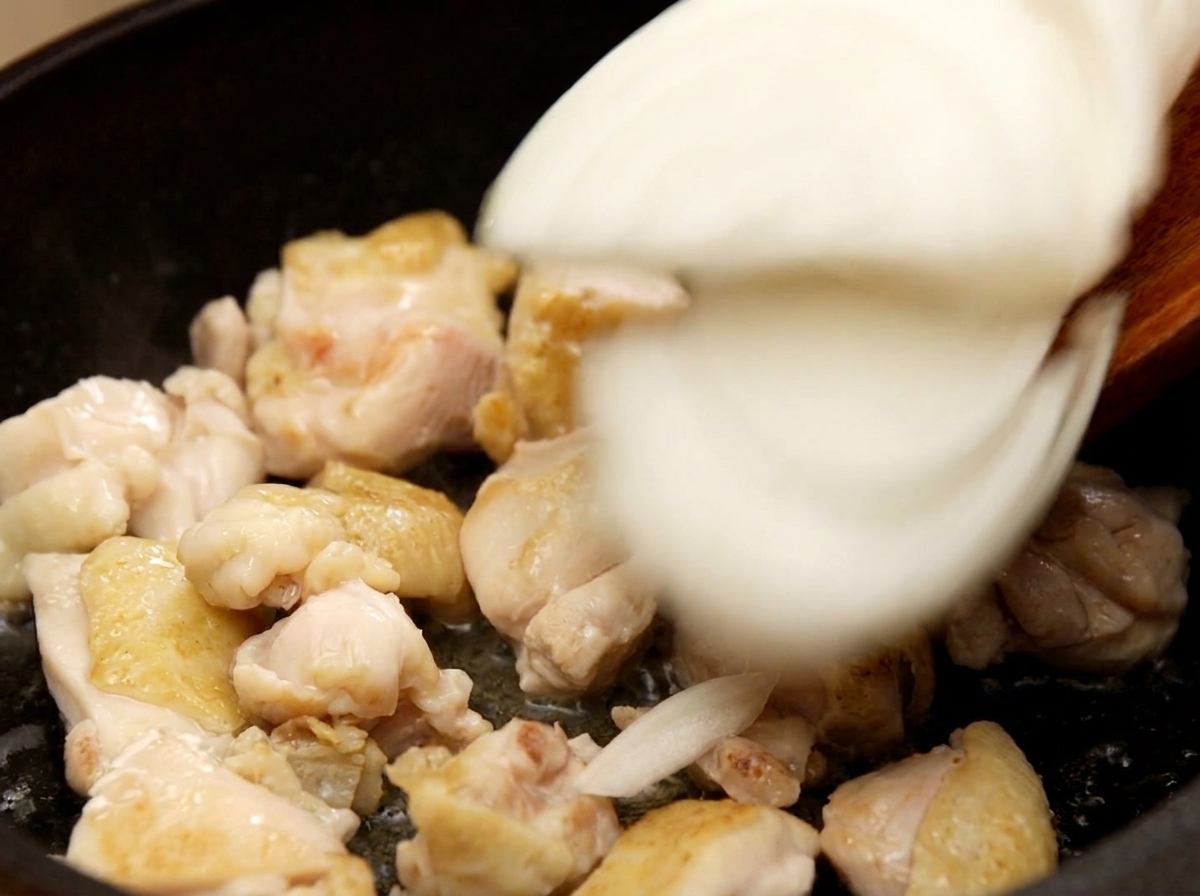 Adding sliced white onions into a frying pan with sautéed chicken pieces and stirring them together.