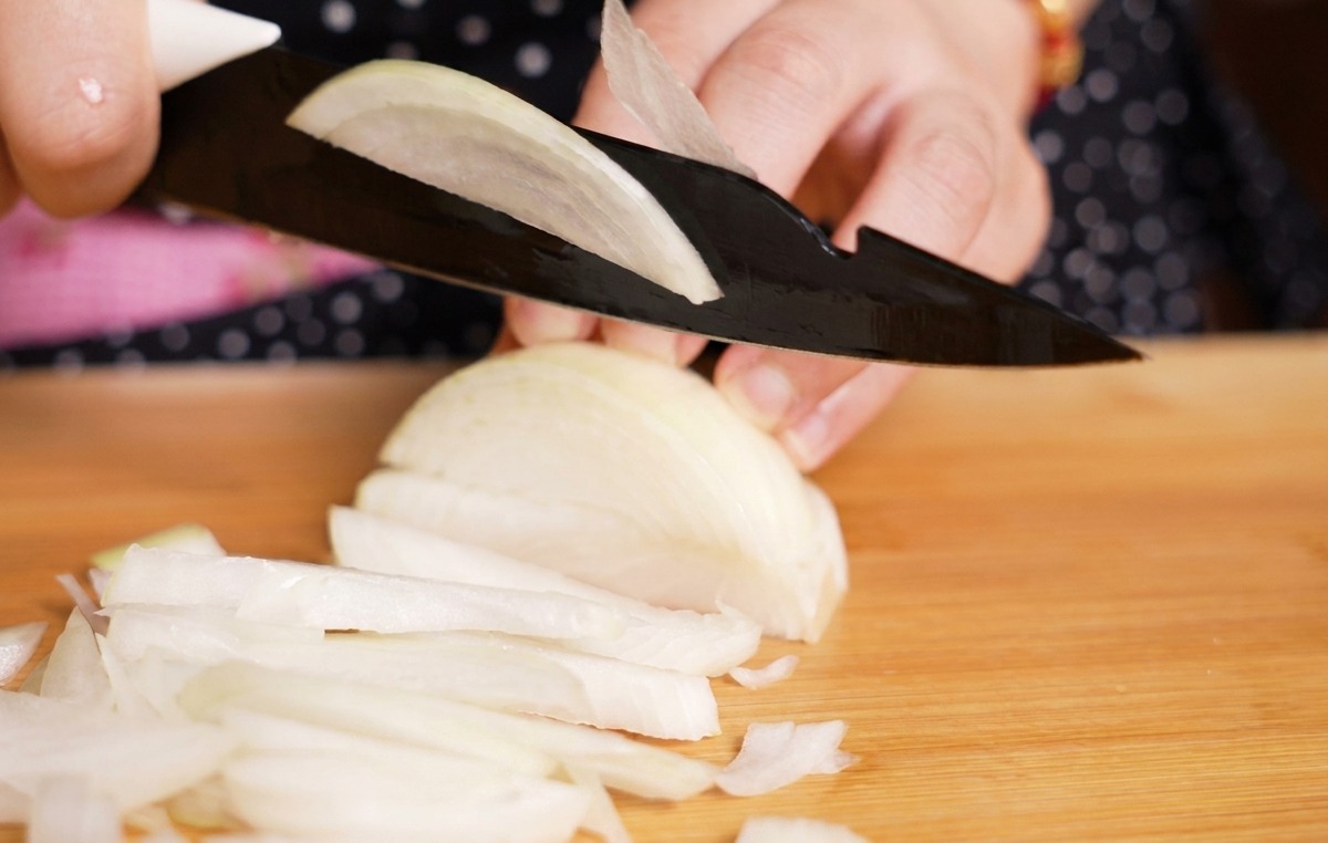 A person using a black knife to slice a white onion into thin strips on a light-colored wooden cutting board.