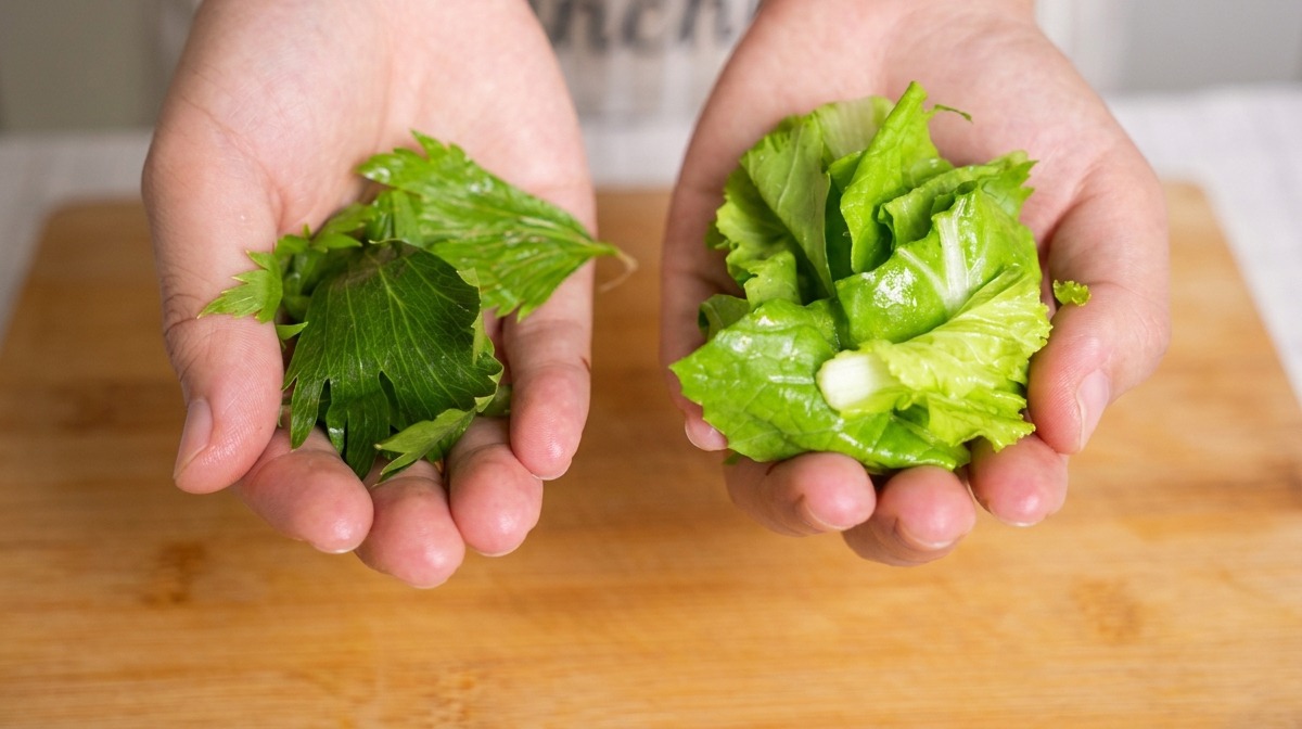 Two hands holding separate handfuls of chopped green celery leaves and baby bok choy over a bamboo board.