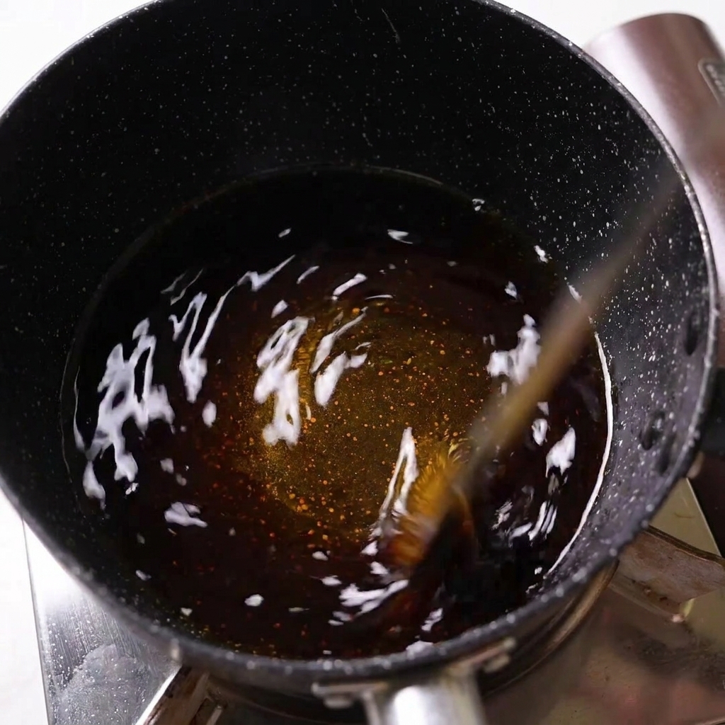 Dark seasoning liquid being stirred in a black speckled saucepan on a stove.