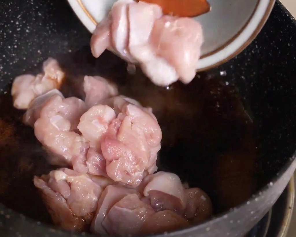 Raw sliced chicken pieces being added from a bowl into a pan of dark simmering sauce.