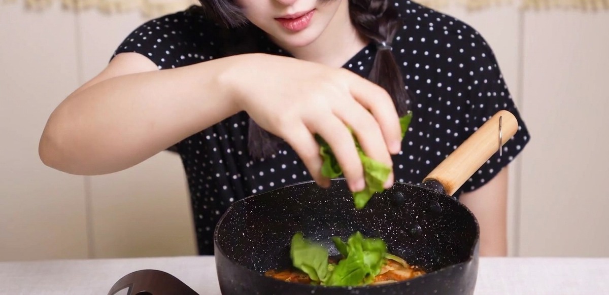 Fresh green vegetables being sprinkled by hand into a pan of simmering chicken, onions, and eggs in a savory dashi sauce.