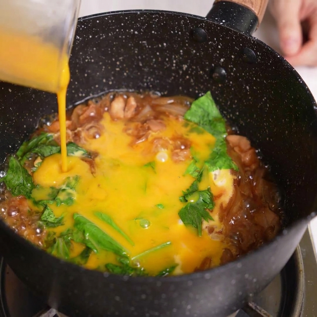Pouring the final portion of beaten egg from a measuring cup over the simmering Oyakodon ingredients.