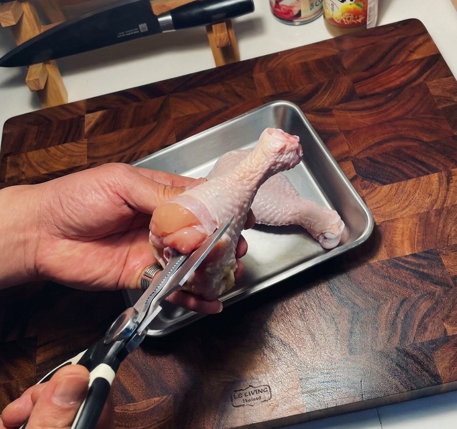 A pair of kitchen shears removing the bone from a raw chicken thigh held over a metal tray.