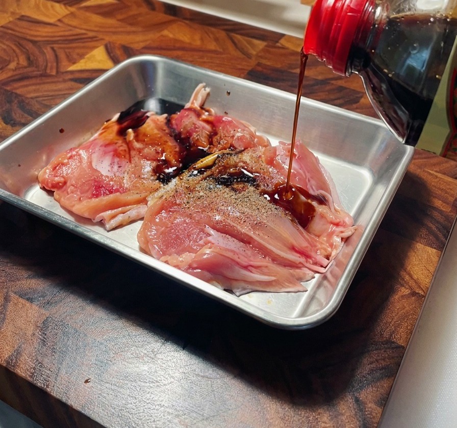 Soy sauce being poured from a bottle onto raw, peppered chicken thighs in a stainless steel tray.