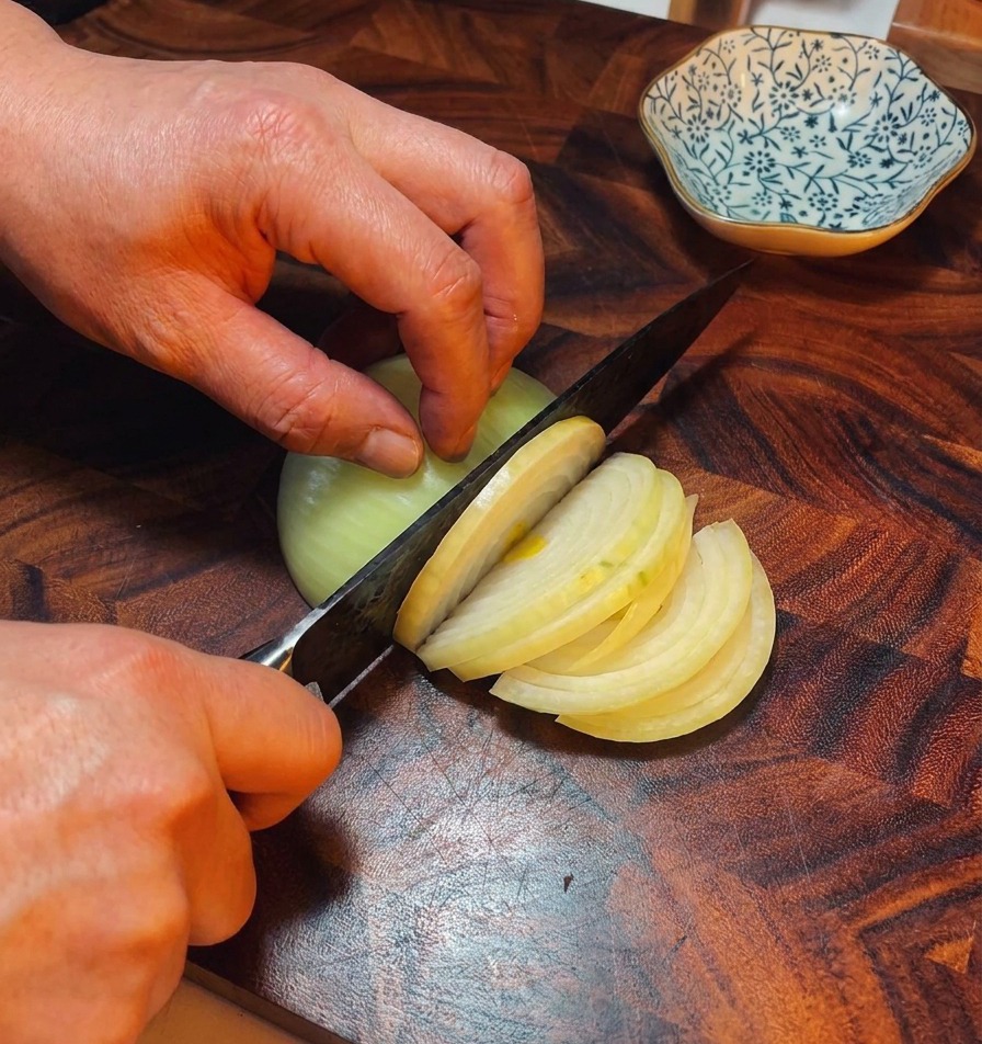 A chefs hands using a knife to slice a yellow onion into thin strips on a dark wood cutting board.