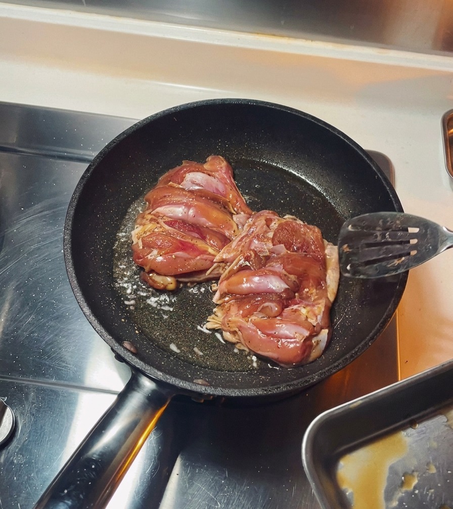 Two seasoned chicken thighs frying skin-side down in a black pan until the skin is browned.