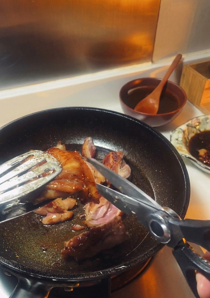 A pair of kitchen shears cutting into a browned chicken thigh being held by metal tongs inside a frying pan.