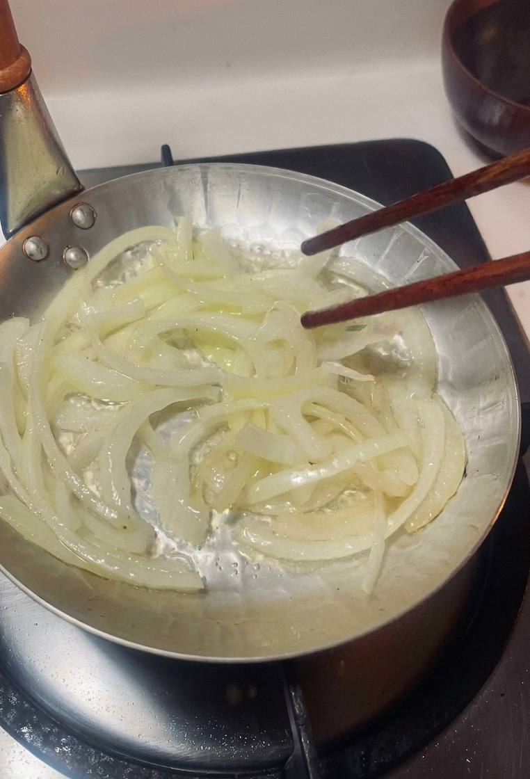 Thinly sliced onions being stirred with wooden chopsticks in a small silver pan over a gas burner.