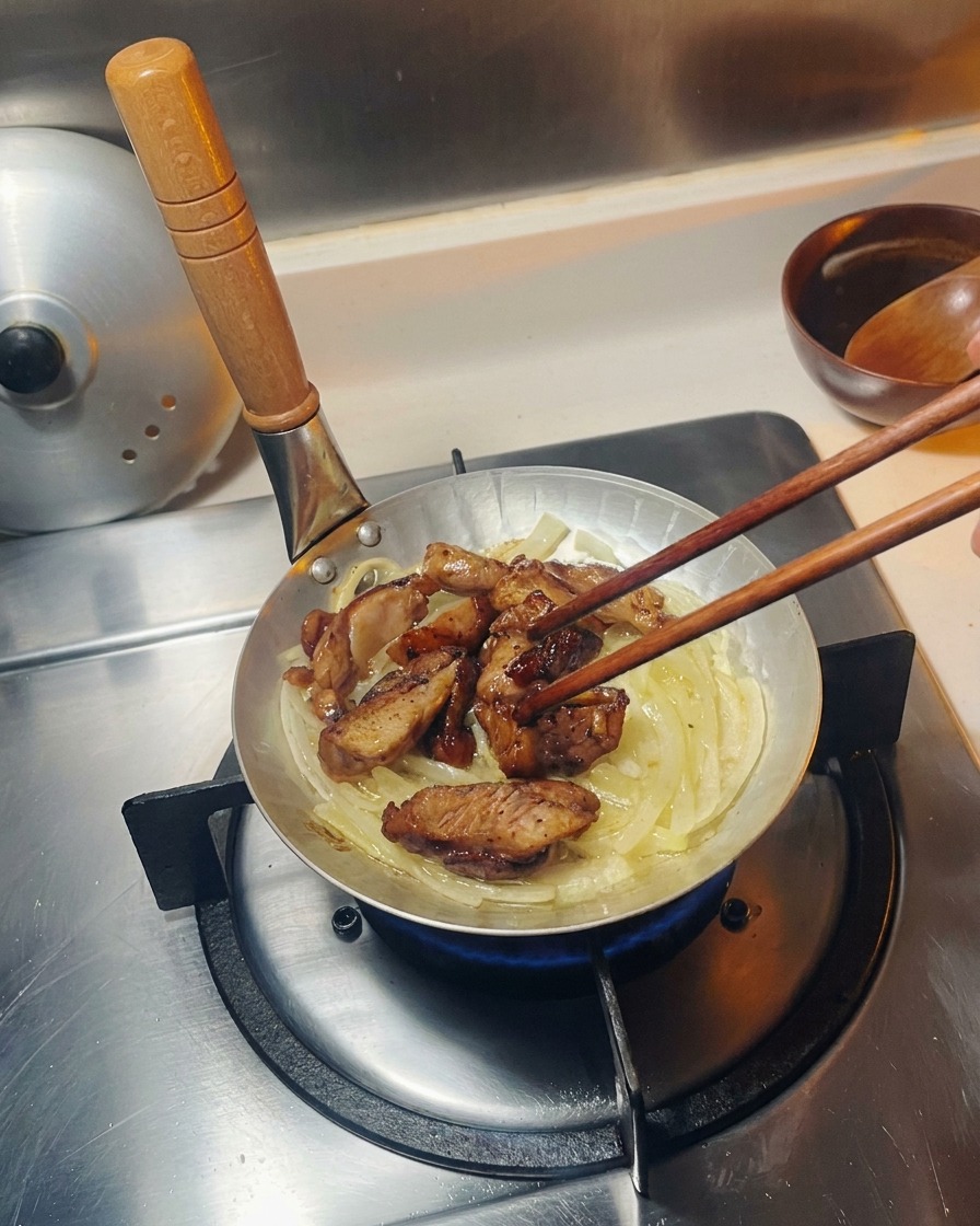 Fried chicken pieces being placed on top of a bed of translucent sautéed onions in a small silver cooking pan.