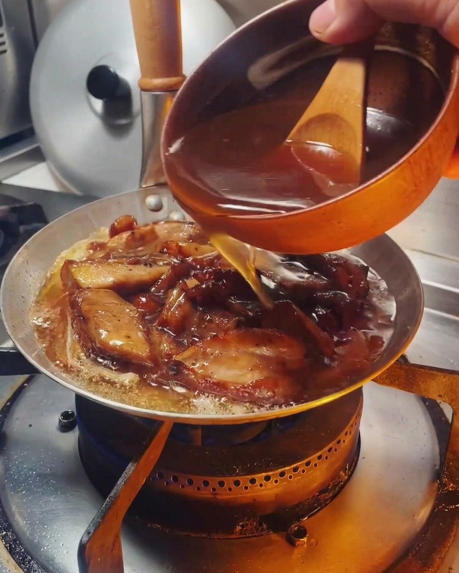 A savory brown sauce being poured from a small wooden-handled bowl into a pan containing chicken and onions.