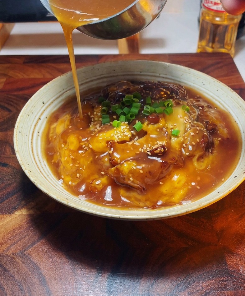 A close up view of a hand using a small metal ladle to pour a golden brown savory sauce over a plated bowl of chicken and egg garnished with green scallions.