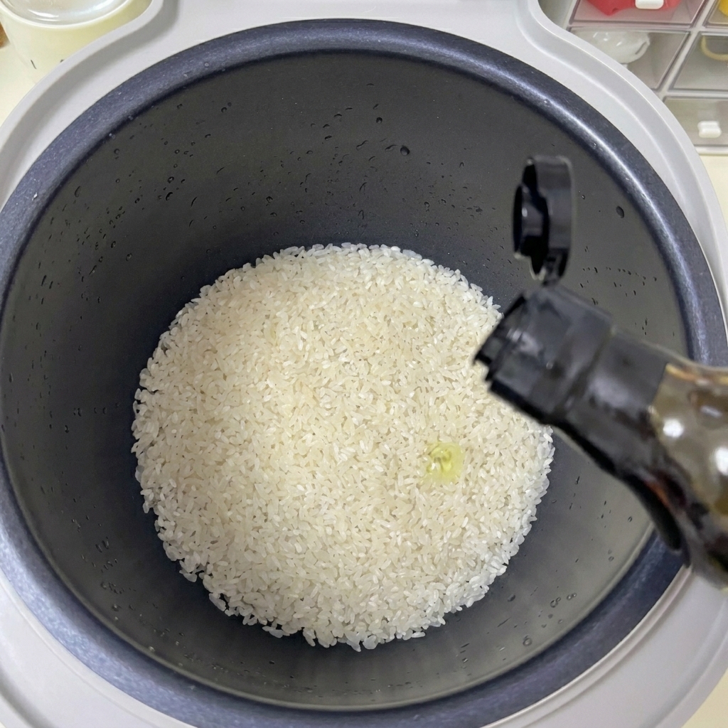 A hand pouring a stream of clear oil from a dark bottle into a rice cooker pot filled with white rice.