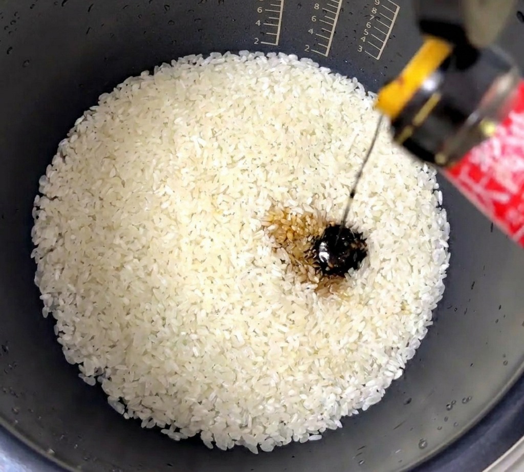 A stream of dark, thick oyster sauce being poured into a rice cooker pot filled with washed white rice.