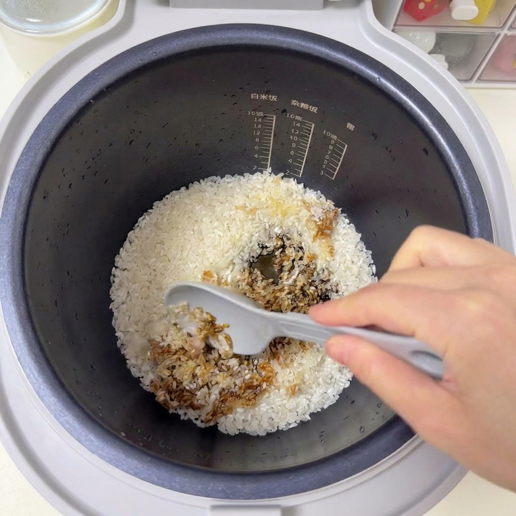 A grey spatula stirring white rice and brown sauces together inside a rice cooker pot.