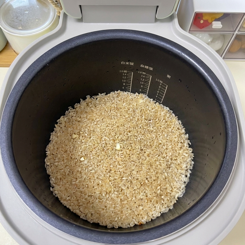Seasoned light brown rice flattened evenly at the bottom of a black rice cooker pot.