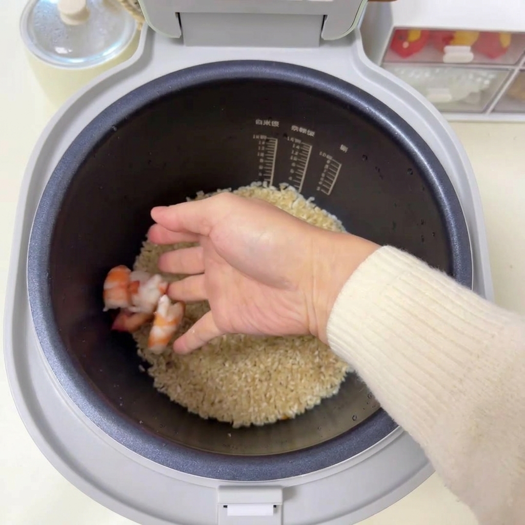 A hand placing several raw pink prawns onto a bed of seasoned rice inside a rice cooker liner.