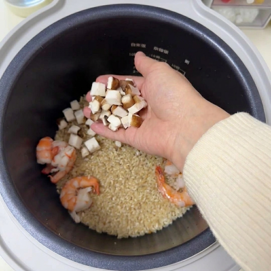 Diced pieces of brown shiitake mushrooms being held in a hand over a rice cooker containing rice and prawns.