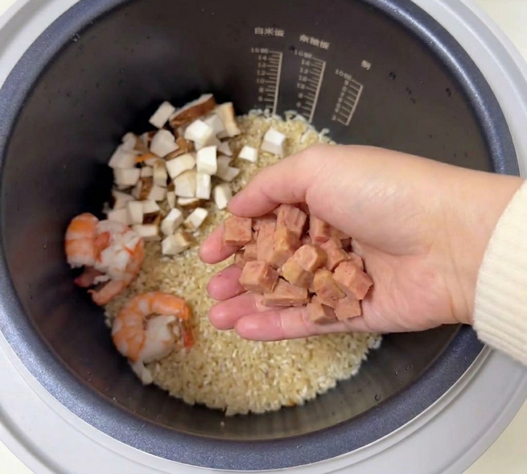 A hand holding a handful of diced pink luncheon meat over a rice cooker pot containing seasoned rice and prawns.