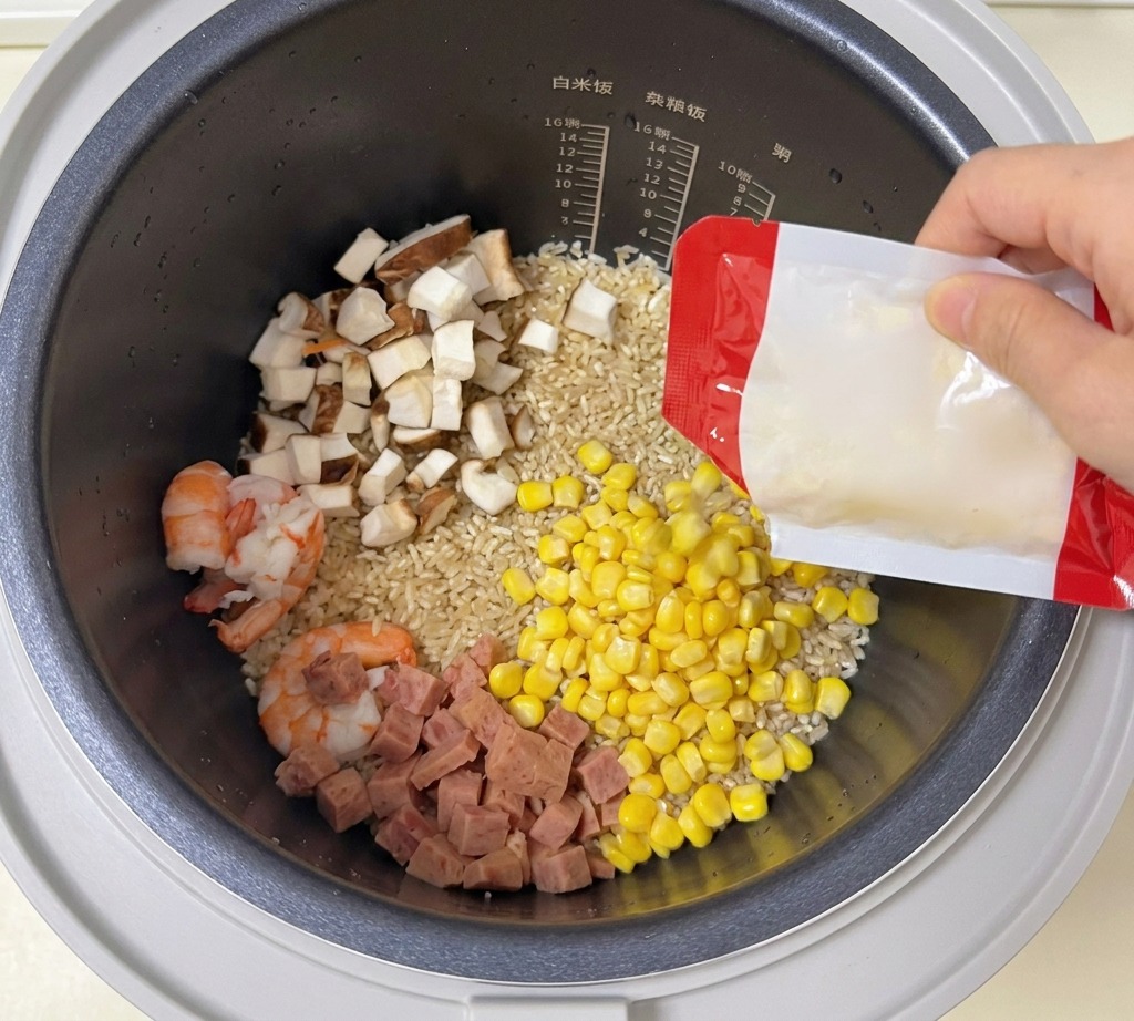 Sweet corn kernels being poured from a packet into a rice cooker pot containing rice, prawns, mushrooms, and luncheon meat.