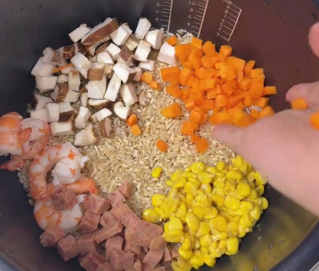 A colorful arrangement of diced carrots, corn, luncheon meat, mushrooms, and prawns layered on top of rice in a pot.