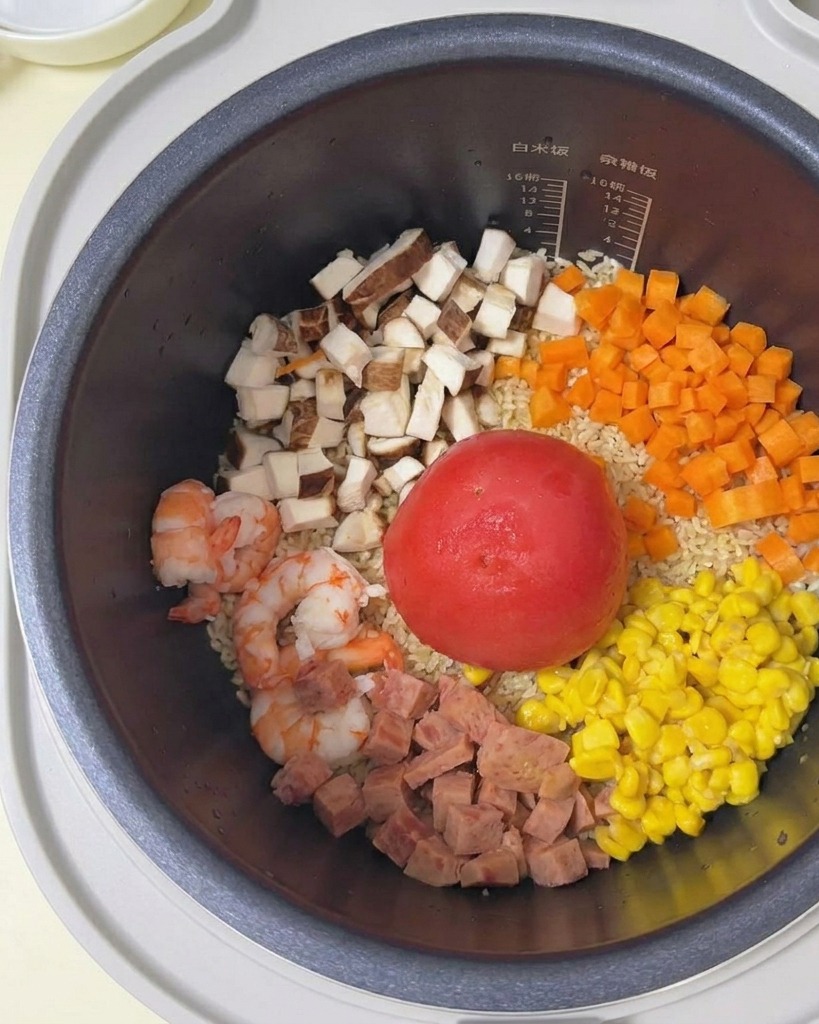 Clear water being poured from a white bowl into a rice cooker pot filled with rice, a whole tomato, and various diced ingredients.