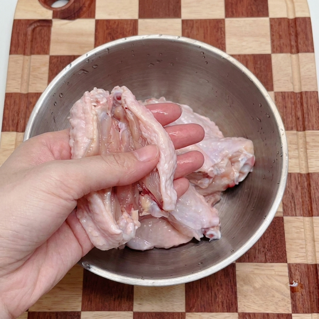 A pair of hands using silver kitchen shears to snip raw chicken wings into two pieces inside a stainless steel mixing bowl.