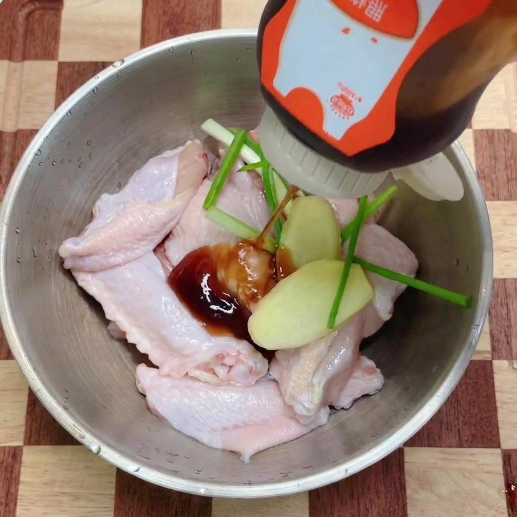 Dark teriyaki sauce being drizzled from a squeeze bottle onto a bowl of raw chicken wings garnished with fresh aromatics.