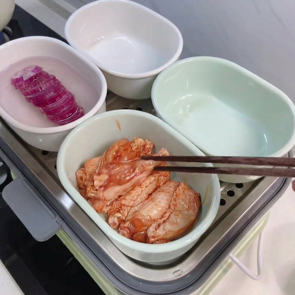 Wooden chopsticks arranging marinated chicken wings in a white bowl, next to another bowl containing rice and purple sweet potato slices.