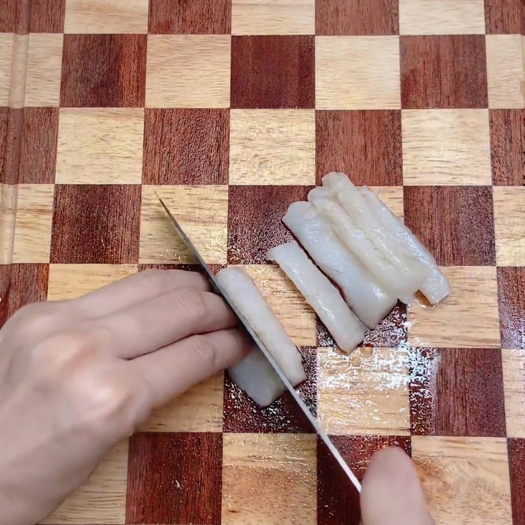A kitchen knife slicing a white cod fillet into even strips on a checkered wooden cutting board.
