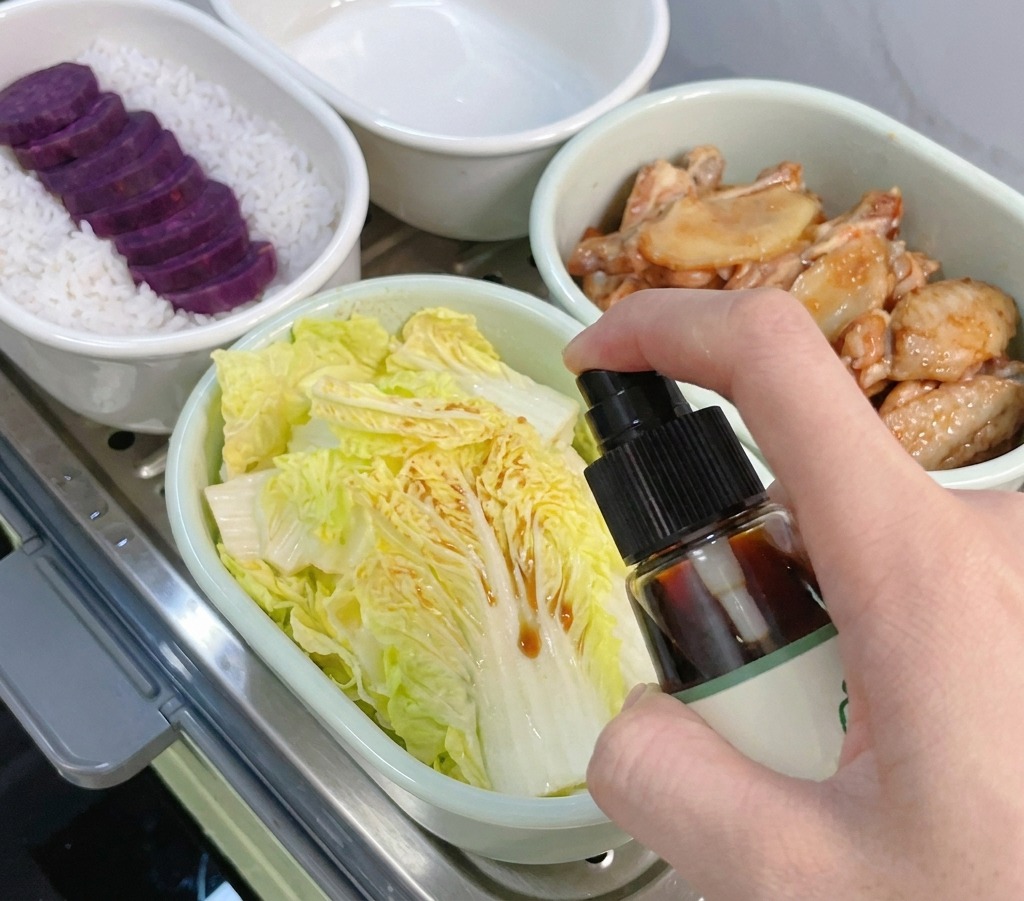 A hand holding a spray bottle and misting dark soy sauce over light green baby cabbage leaves in a white bowl.