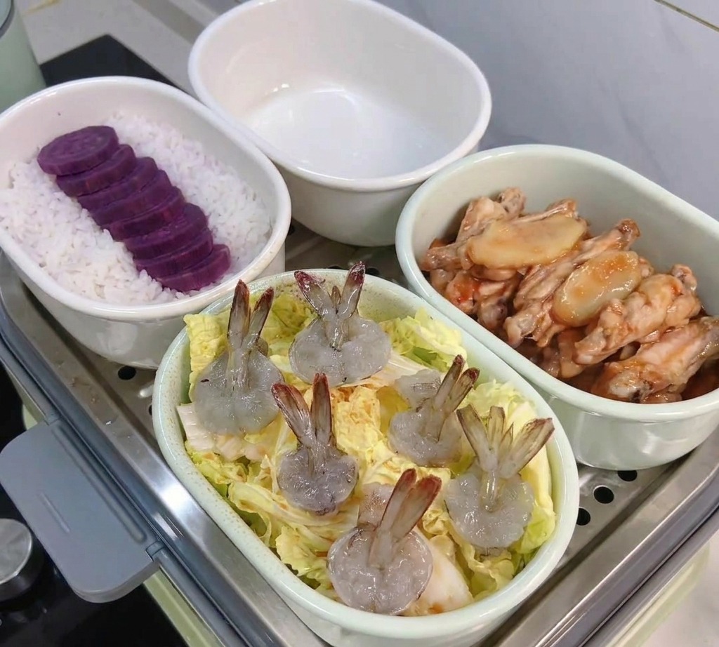 A white oval dish containing layered baby cabbage leaves topped with several raw, greyish-pink peeled shrimp, ready for the steamer.