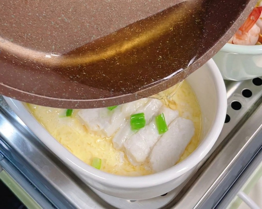 Hot clear oil being poured from a brown frying pan onto chopped scallions and steamed cod fish sitting on top of a yellow egg custard.