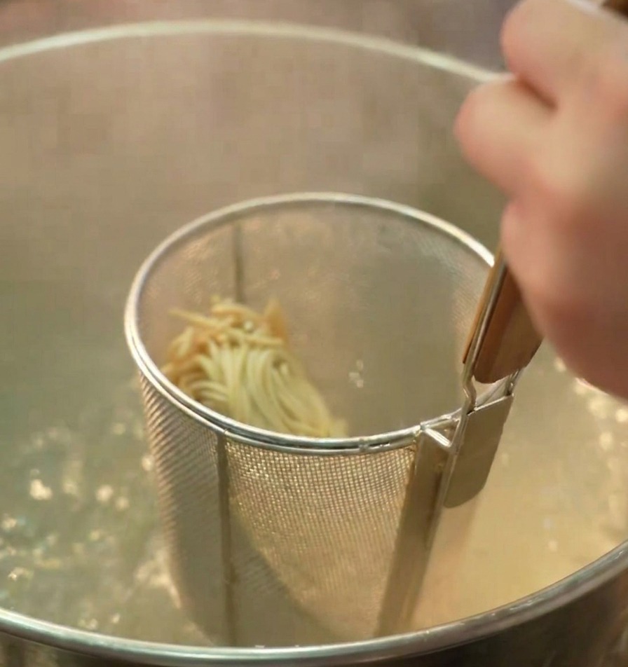 A mesh strainer containing ramen noodles is held inside a deep pot of bubbling boiling water.