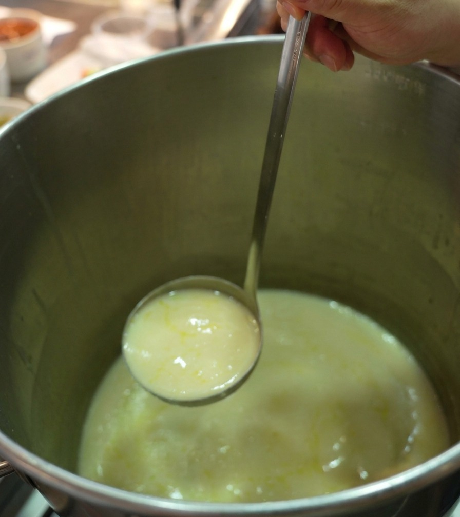 A stainless steel ladle lifting thick, creamy yellow chicken broth from a large professional stockpot.