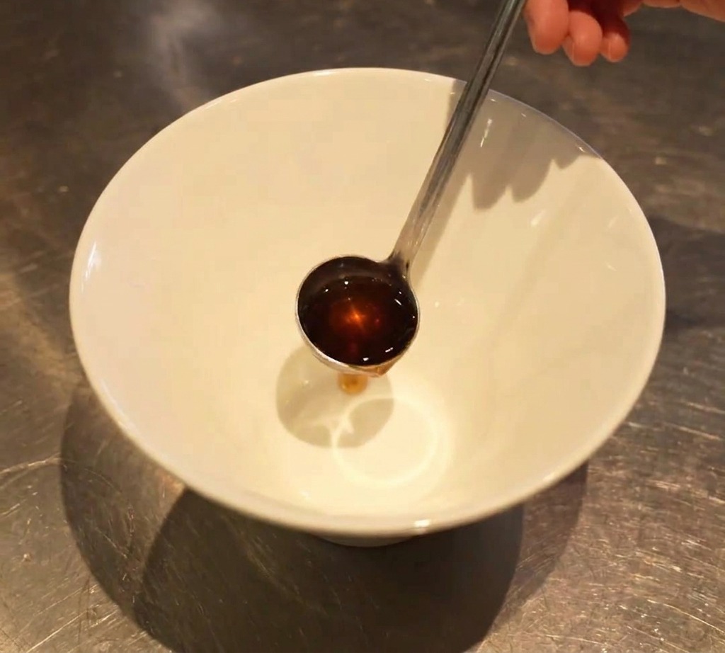 A small metal ladle pouring a dark liquid seasoning into the base of an empty white bowl.