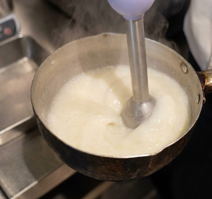 An immersion blender submerged in a pan of yellow liquid, creating a thick, white, frothy foam on the surface.