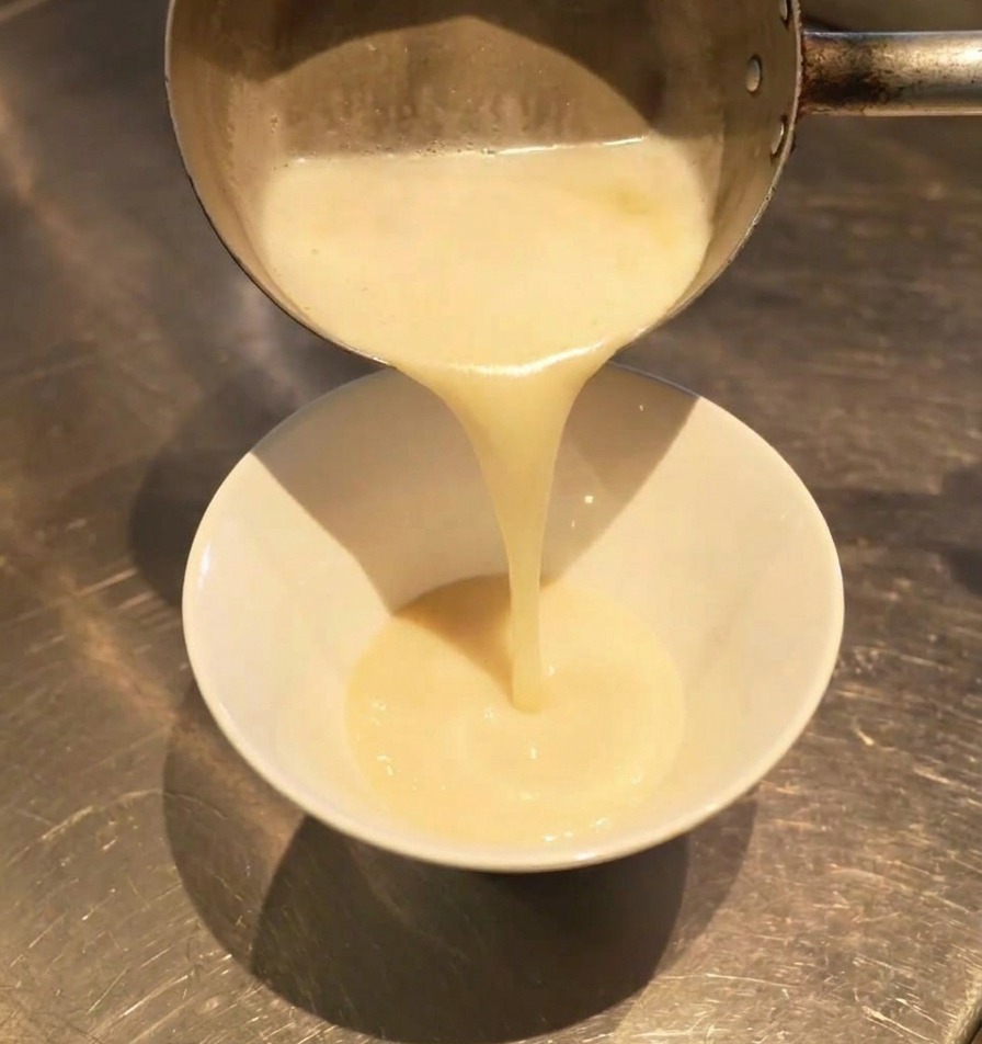 Thick, milky white broth pouring from a saucepan into a white bowl, blending with a dark seasoning at the bottom.