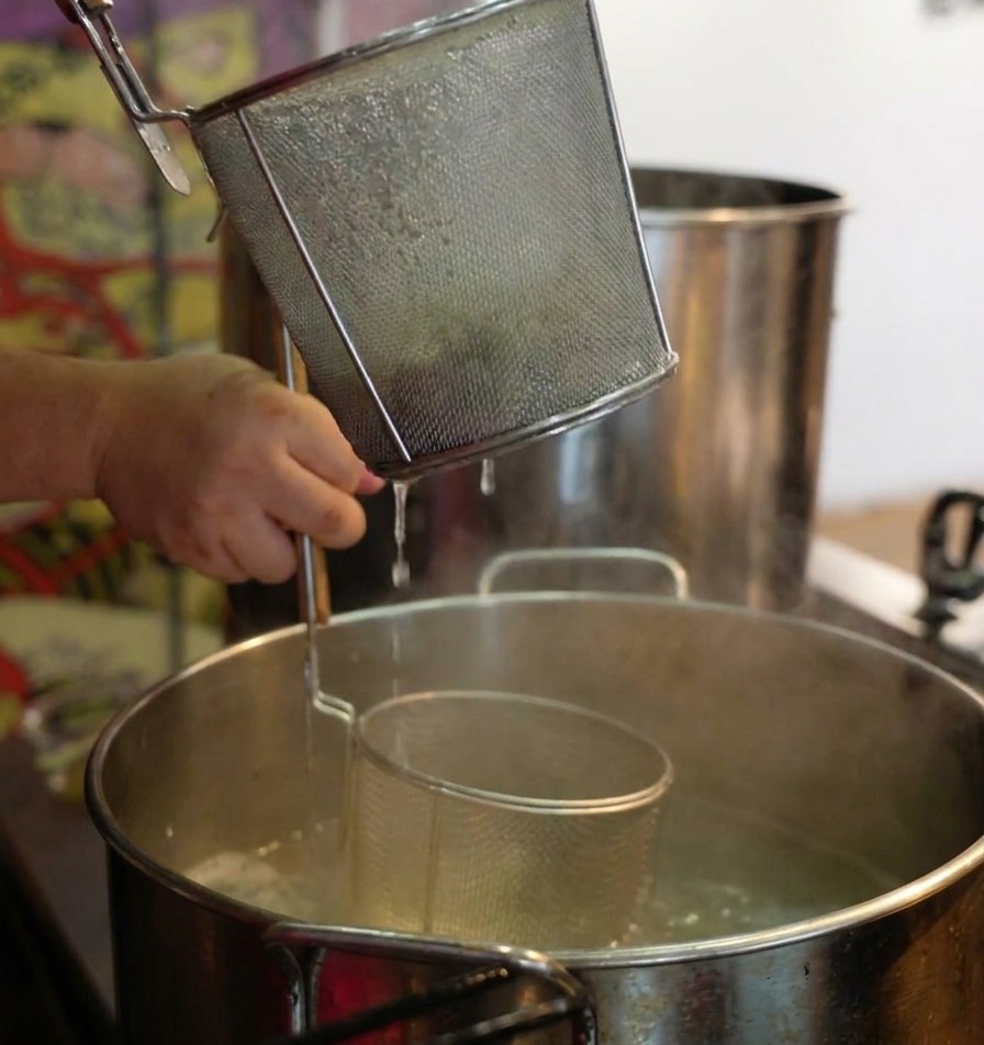 Hands holding a tall wire mesh noodle strainer over a pot of steaming water, shaking it to drain cooked ramen noodles.