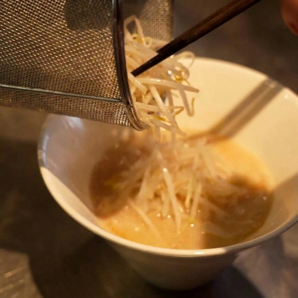 A small wire strainer pouring a pile of white bean sprouts onto a bowl of ramen noodles and broth.