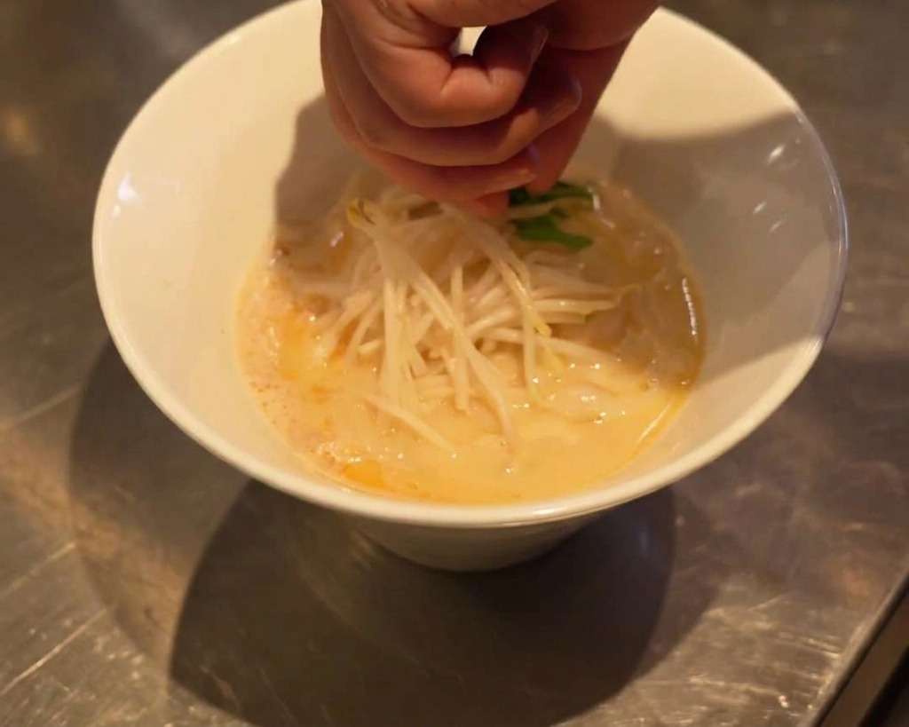 A hand carefully placing fresh green spinach leaves on top of blanched bean sprouts in a white ramen bowl.