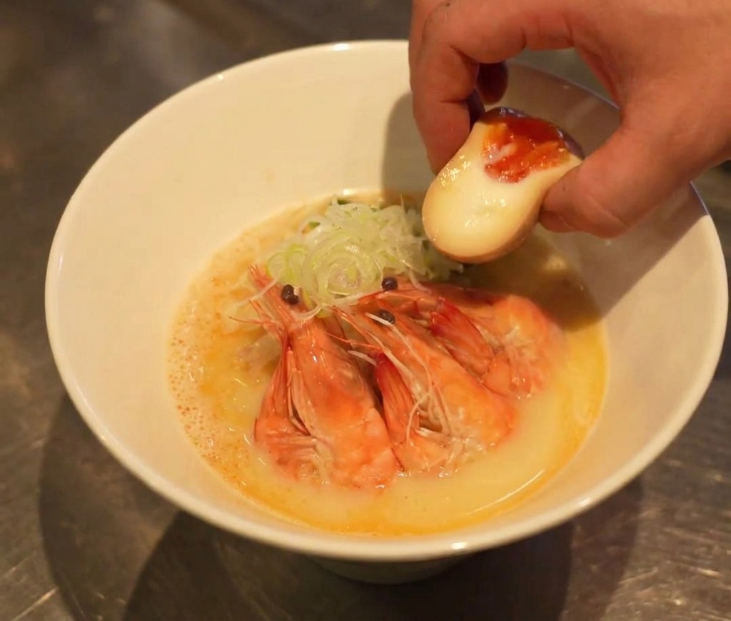 A hand placing a halved soft-boiled egg with a translucent, runny yolk into a nearly assembled bowl of ramen.
