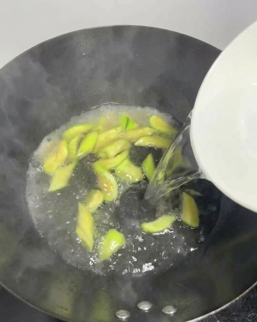 Clear water being poured from a white bowl into a bubbling wok with silk melon slices.
