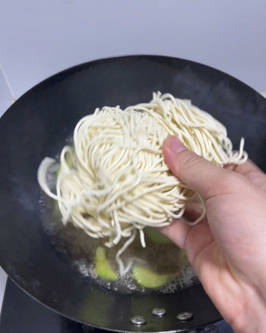 A hand placing a bundle of fresh ramen noodles into a wok of boiling broth.
