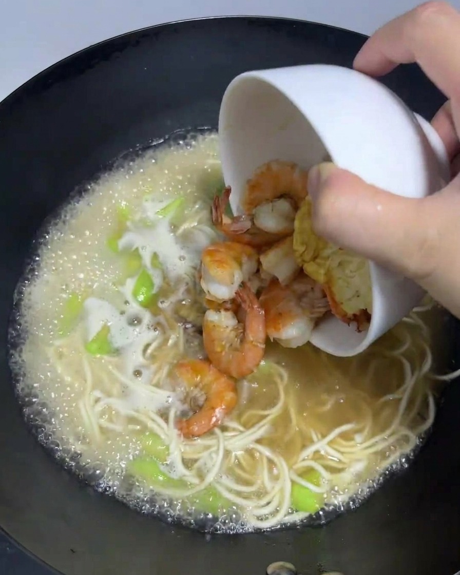 Cooked shrimp and a fried egg being poured from a bowl back into a simmering wok of ramen noodles.