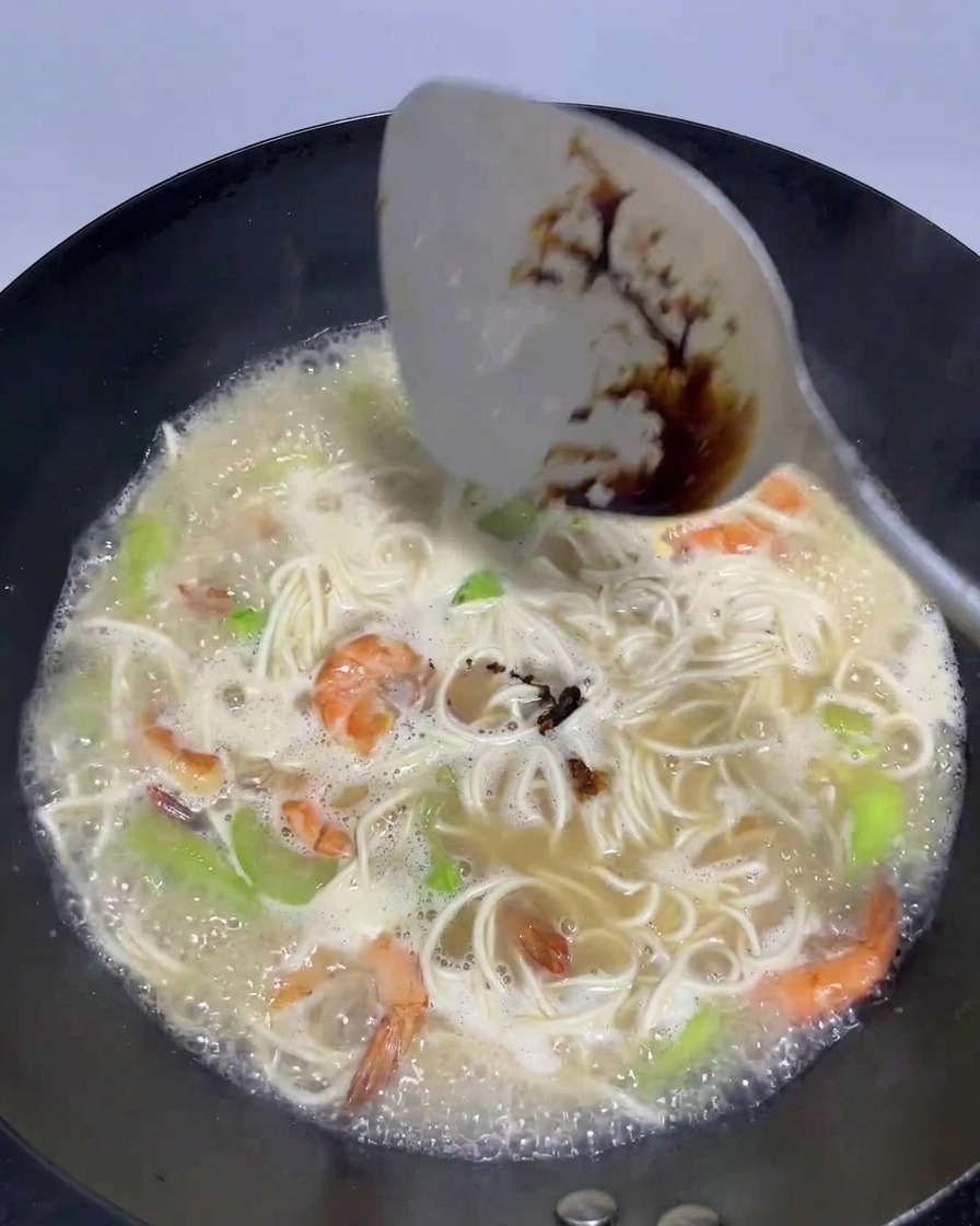 A spatula pouring a splash of dark soy sauce into a wok filled with bubbling broth, noodles, shrimp, and silk melon.